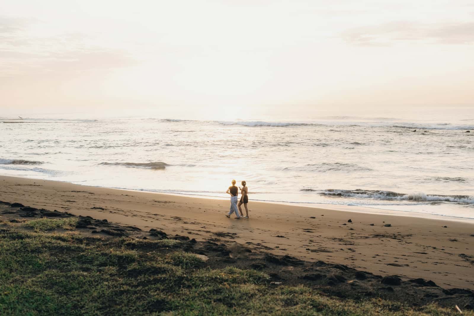 Two people walk side by side along the water’s edge of a wide sandy beach, under a pale sky with the sun low over the sea.