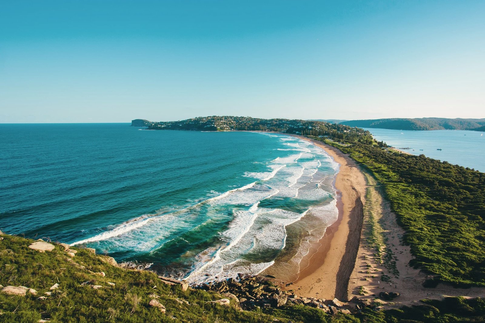 A long, narrow beach seen from high above, with surf on one side and calm water on the other, running to a green headland.