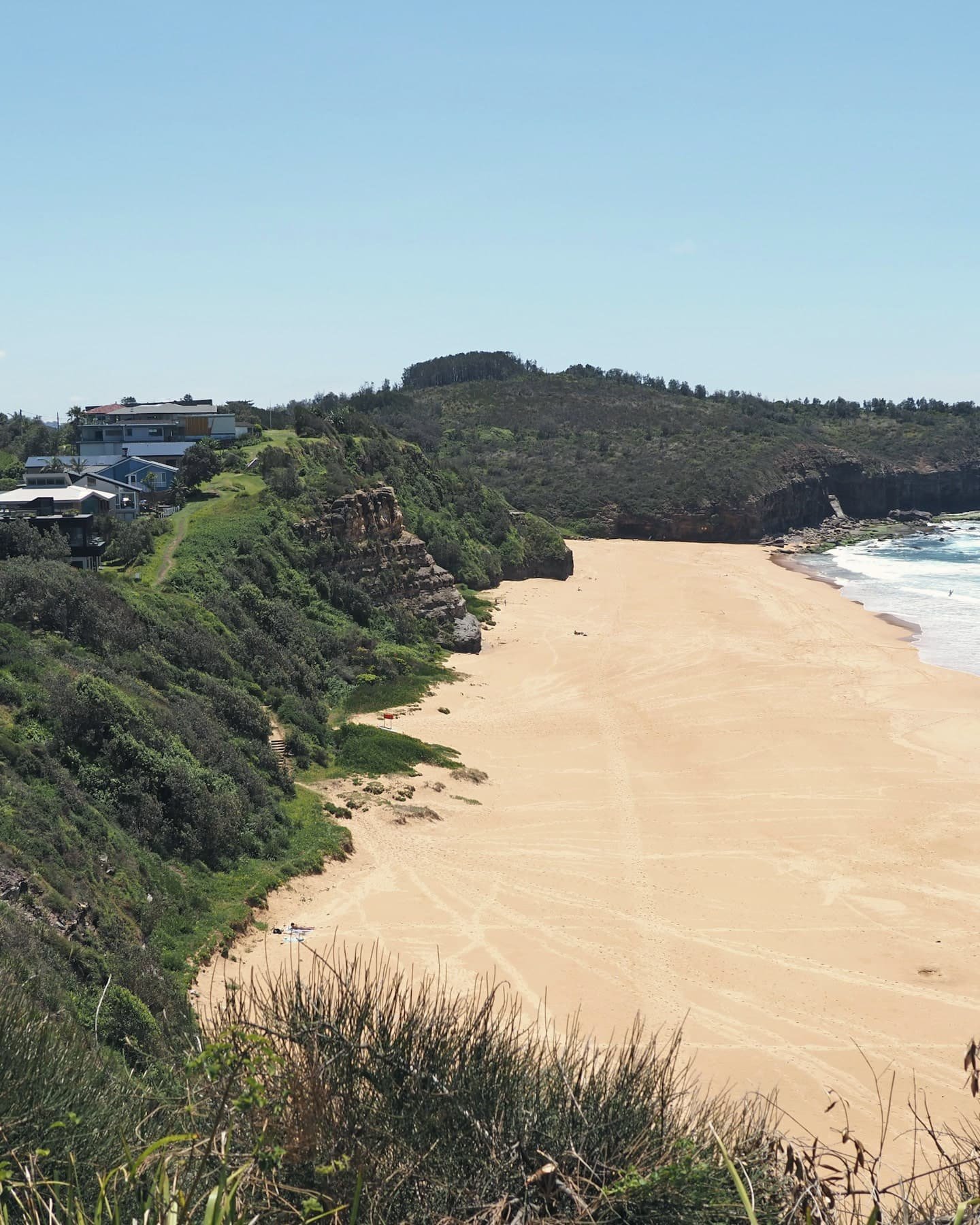 A sandy beach curves below a steep green hillside, with a dark headland and white surf beyond.