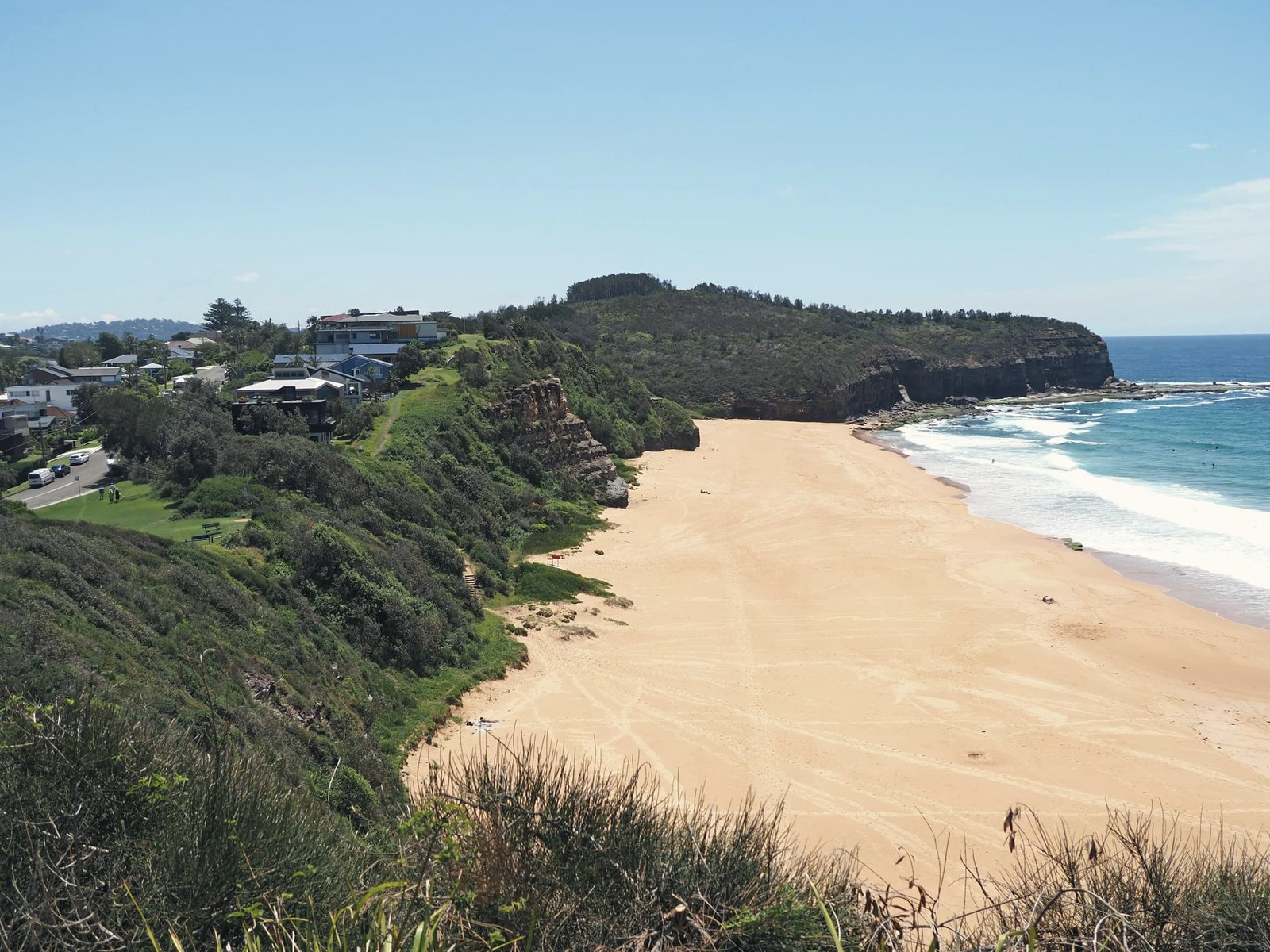A sandy beach curves below a steep green hillside, with a dark headland and white surf beyond.