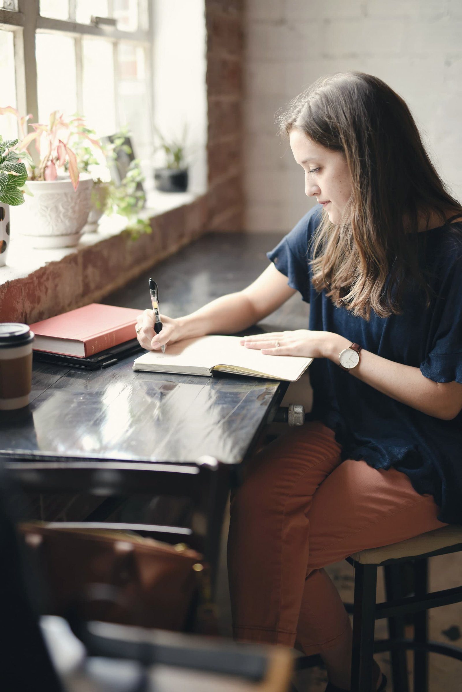 A woman writes in a notebook at a timber table by a sunny window, with plants on the sill and a coffee beside her.