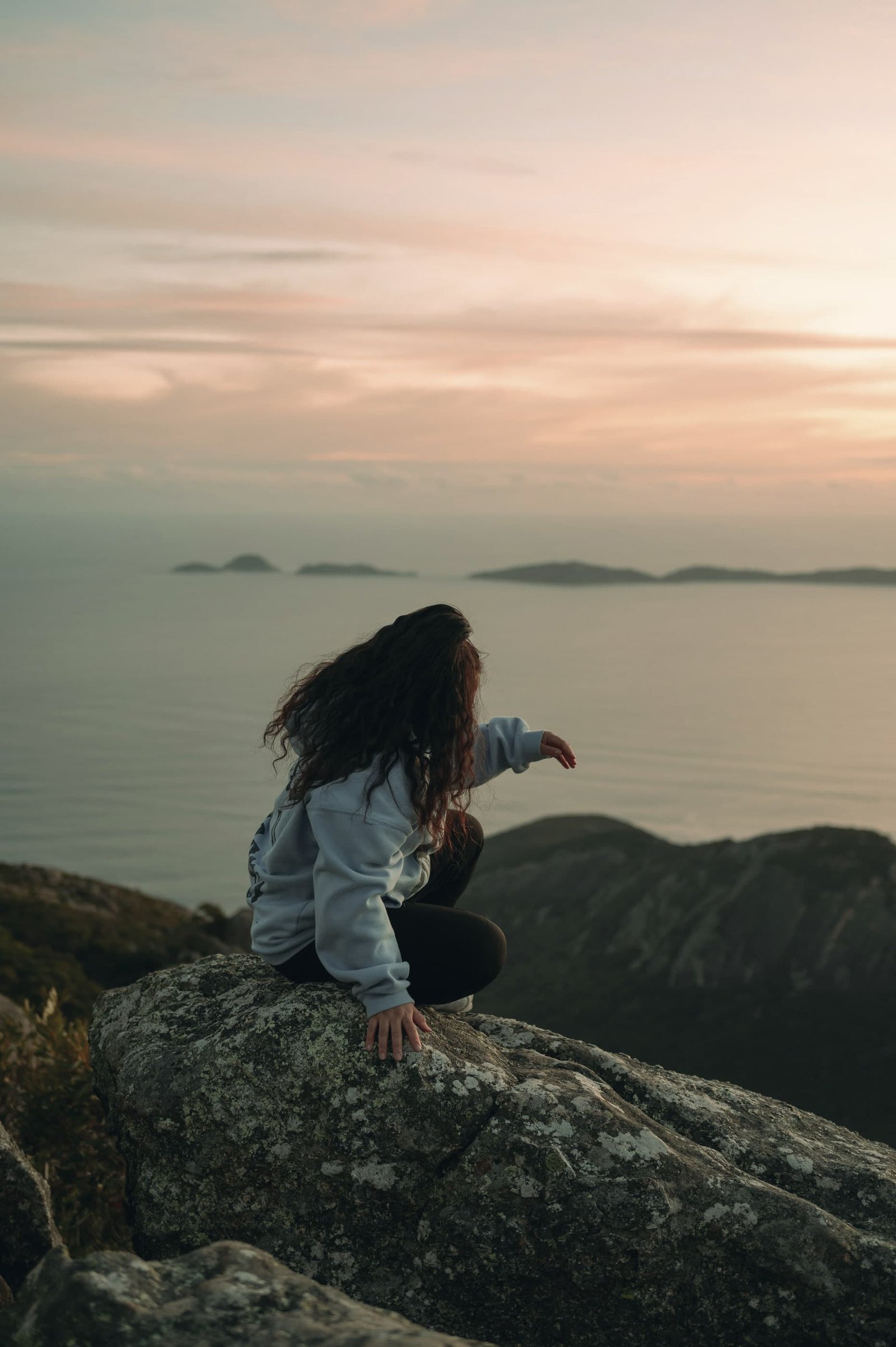 A woman sits on a rock above a calm sea at dusk, looking out towards distant islands under a soft pink sky.